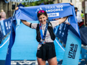 Trail 100 Andorra race recap A woman with dark hair in shorts, t-shirt, and black hydration vest stands at the finish line. She has a medal round her neck and is holding the blue finish tape above her head, smiling.