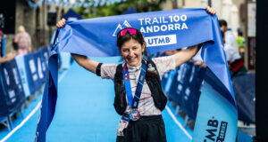 A woman with dark hair in shorts, t-shirt, and black hydration vest stands at the finish line. She has a medal round her neck and is holding the blue finish tape above her head, smiling.