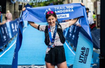 A woman with dark hair in shorts, t-shirt, and black hydration vest stands at the finish line. She has a medal round her neck and is holding the blue finish tape above her head, smiling.