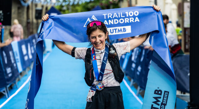 Trail 100 Andorra race recap A woman with dark hair in shorts, t-shirt, and black hydration vest stands at the finish line. She has a medal round her neck and is holding the blue finish tape above her head, smiling.