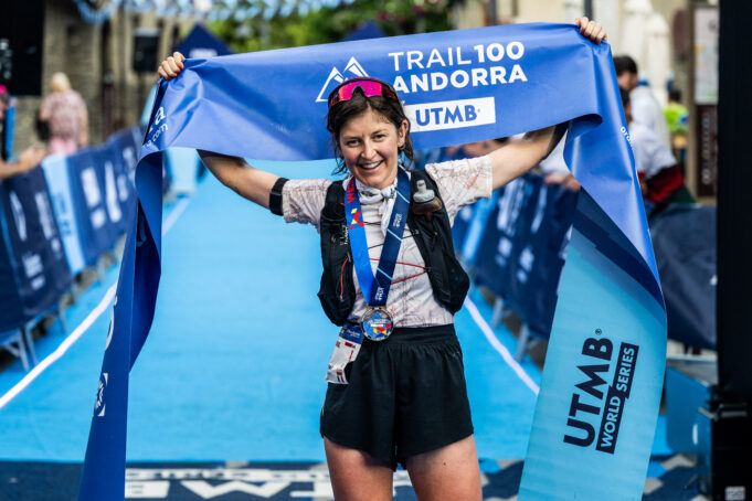 Trail Andorra 105k winner women A woman with dark hair in shorts, t-shirt, and black hydration vest stands at the finish line. She has a medal round her neck and is holding the blue finish tape above her head, smiling.