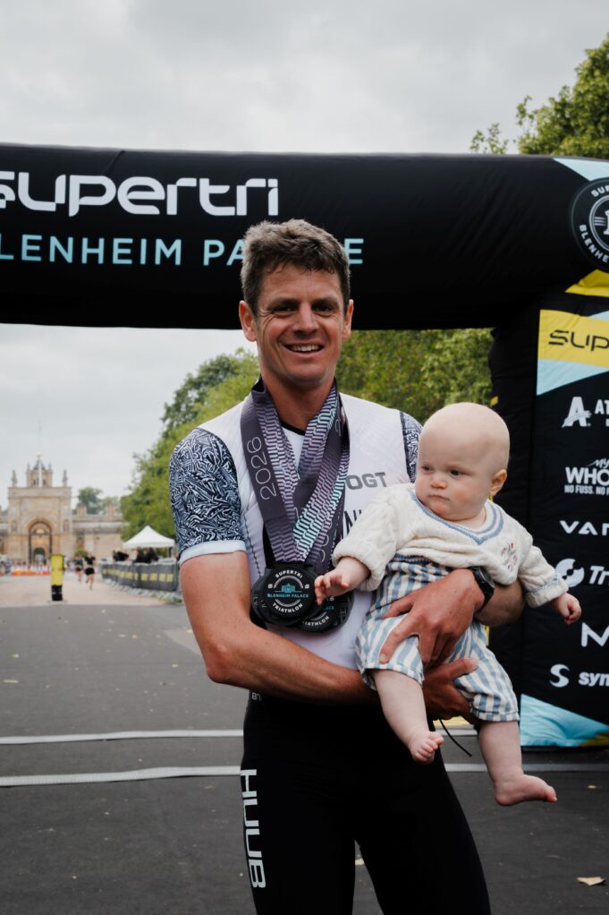 Jonny Brownlee Blenheim Palace Jonny Brownlee stands at the finish line with a medal around his neck, holding his baby