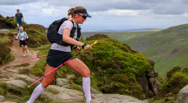 A woman wearing shorts, tshirt, race pack and carrying poles traverses rocky slabs.