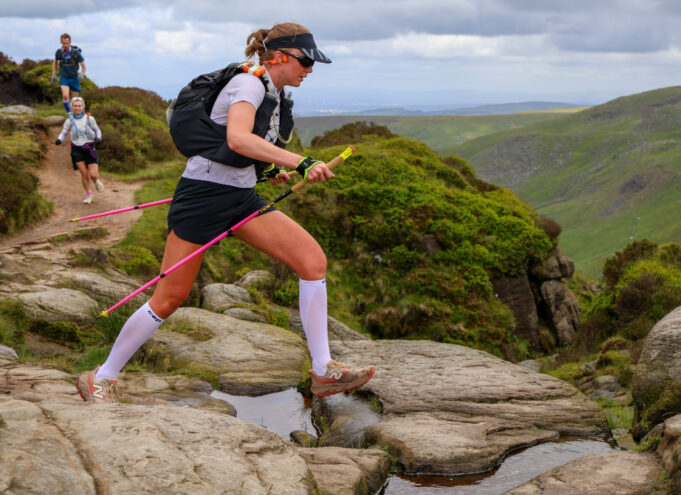 A woman wearing shorts, tshirt, race pack and carrying poles traverses rocky slabs.