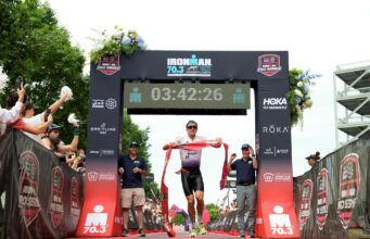 Male triathlete runs under finish gantry holding red tape
