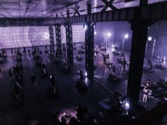 A dark warehouse lit purple via a screen with psychedilic images. The warehouse is full of treadmills