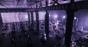 A dark warehouse lit purple via a screen with psychedilic images. The warehouse is full of treadmills