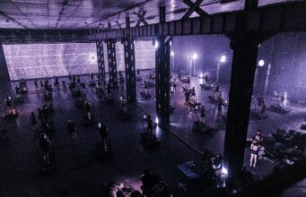 24 hours in the Treadmill Factory A dark warehouse lit purple via a screen with psychedilic images. The warehouse is full of treadmills