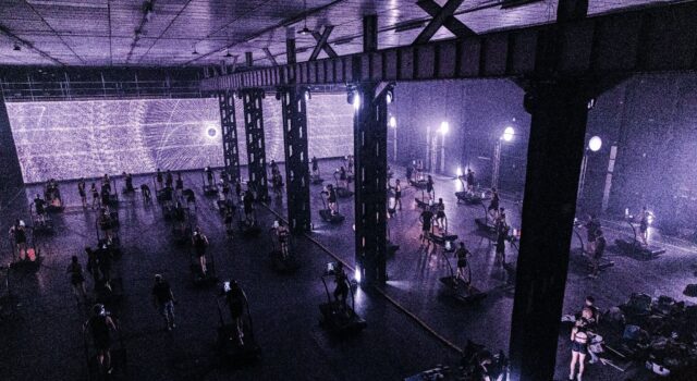 24 hours in the Treadmill Factory A dark warehouse lit purple via a screen with psychedilic images. The warehouse is full of treadmills