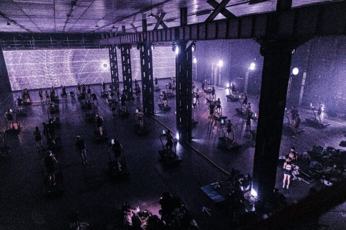 ARC Treadmill A dark warehouse lit purple via a screen with psychedilic images. The warehouse is full of treadmills