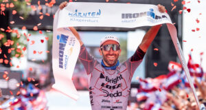 Male triathlete in sunglasses liftes finish tape above head as confetti falls around him.
