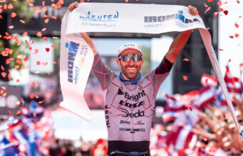 Male triathlete in sunglasses liftes finish tape above head as confetti falls around him.