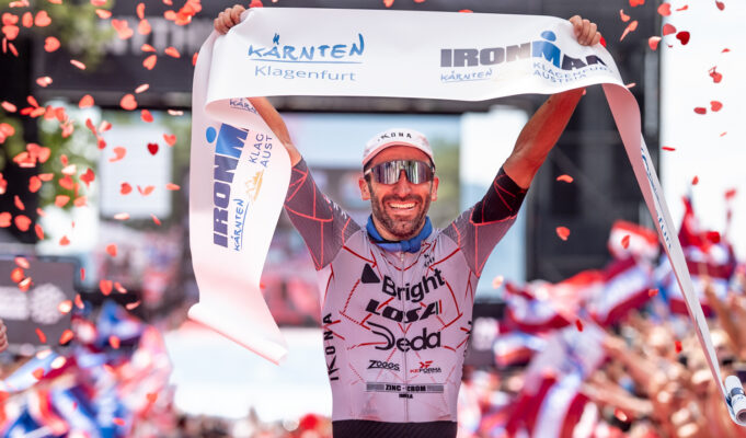 Mattia Ceccarelli (ITA) Takes His First Professional IRONMAN Win at IRONMAN Kärnten-Klagenfurt, Austria Male triathlete in sunglasses liftes finish tape above head as confetti falls around him.