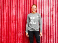 Mel Bound awarded OBE for women’s community sport Smiling woman in front of red background, wearing grey sweatshirt saying I RUN THE SHOW