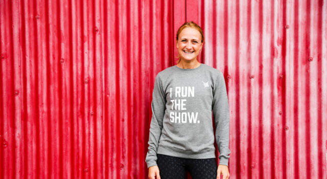 Smiling woman in front of red background, wearing grey sweatshirt saying I RUN THE SHOW