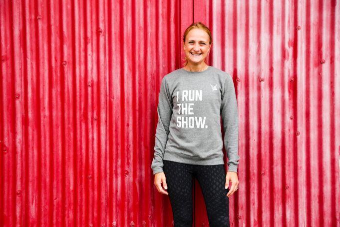 Mel Bound Smiling woman in front of red background, wearing grey sweatshirt saying I RUN THE SHOW