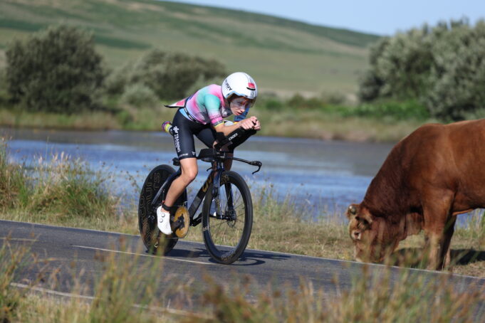 IRONMAN 70.3 Swansea Pro Series Woman on bike cycles near lake past brown cow