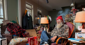 An elderly man wering running shoes, jeans, plaid shirt and red beany hat sits in an armchair in a room full of stuff