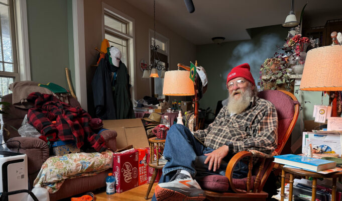 An elderly man wering running shoes, jeans, plaid shirt and red beany hat sits in an armchair in a room full of stuff