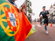 A male triathlete in a tri suit runs past a Portuguese flag