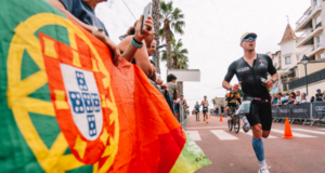 A male triathlete in a tri suit runs past a Portuguese flag