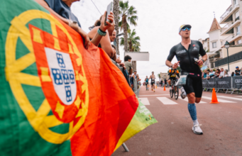 A male triathlete in a tri suit runs past a Portuguese flag
