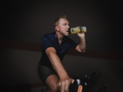 Blond man on a static bike drinks out of a sports bottle in a dark room.