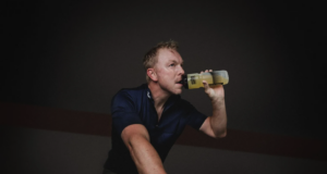 Blond man on a static bike drinks out of a sports bottle in a dark room.