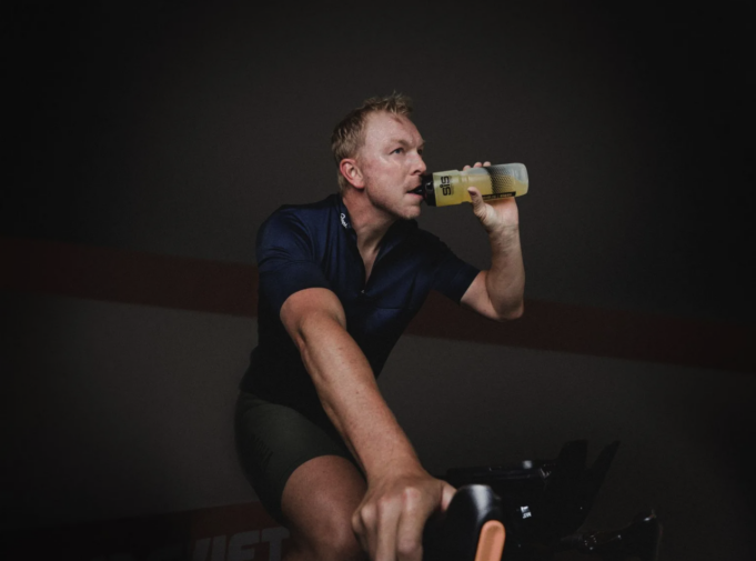 Blond man on a static bike drinks out of a sports bottle in a dark room.