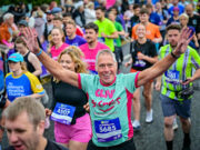 Run For All launches 2027 season tickets A man in a mint green and pink t-shirt is centre of the image. He has his arms raised and is looking at the camera, smiling. Around him is a mass of runners in neon