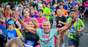 A man in a mint green and pink t-shirt is centre of the image. He has his arms raised and is looking at the camera, smiling. Around him is a mass of runners in neon