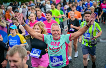 Run For All launches 2027 season tickets A man in a mint green and pink t-shirt is centre of the image. He has his arms raised and is looking at the camera, smiling. Around him is a mass of runners in neon