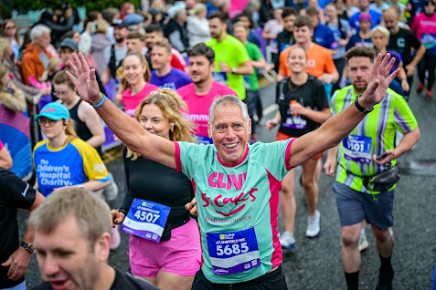 Run For All launches 2027 season tickets A man in a mint green and pink t-shirt is centre of the image. He has his arms raised and is looking at the camera, smiling. Around him is a mass of runners in neon