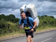 Meet The Bale Man: running races with a 20kg hay bale Man in blue t-shirt and black shorts runs along a road. He is leaning forward with the weight of a 20kg shrink wrapped hay bale which he carries on his back.