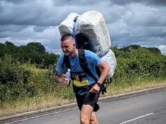 Meet The Bale Man: running races with a 20kg hay bale Man in blue t-shirt and black shorts runs along a road. He is leaning forward with the weight of a 20kg shrink wrapped hay bale which he carries on his back.