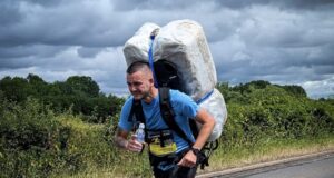 Man in blue t-shirt and black shorts runs along a road. He is leaning forward with the weight of a 20kg shrink wrapped hay bale which he carries on his back.