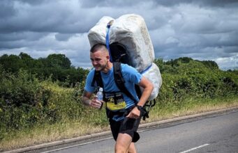 Meet The Bale Man: running races with a 20kg hay bale Man in blue t-shirt and black shorts runs along a road. He is leaning forward with the weight of a 20kg shrink wrapped hay bale which he carries on his back.