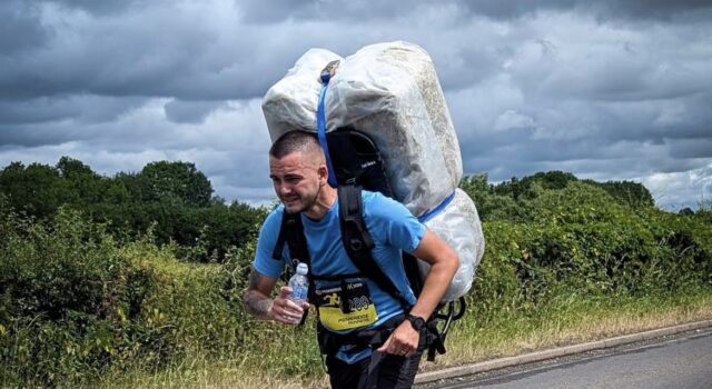 Meet The Bale Man: running races with a 20kg hay bale Man in blue t-shirt and black shorts runs along a road. He is leaning forward with the weight of a 20kg shrink wrapped hay bale which he carries on his back.