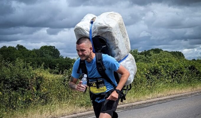Man in blue t-shirt and black shorts runs along a road. He is leaning forward with the weight of a 20kg shrink wrapped hay bale which he carries on his back.