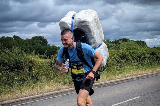 Lee Firmstone the Bale Man Man in blue t-shirt and black shorts runs along a road. He is leaning forward with the weight of a 20kg shrink wrapped hay bale which he carries on his back.