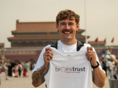 Smiling man in white tshirt and black shorts holds a brainstrust white vest. He stands in front of a Chinese temple