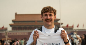 Smiling man in white tshirt and black shorts holds a brainstrust white vest. He stands in front of a Chinese temple