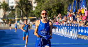 Female athlete in blue crosses finish line. She's holding the blue finish tape in one hand, and yelling in delight