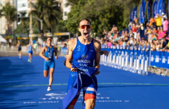 Female athlete in blue crosses finish line. She's holding the blue finish tape in one hand, and yelling in delight