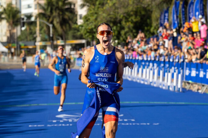 Zuzana Michalickova World Triathlon Female athlete in blue crosses finish line. She's holding the blue finish tape in one hand, and yelling in delight