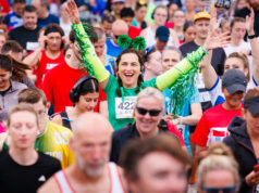 Lady in green in the middle of a crowd of runners. She has her arms outstretched and looks overjoyed to be there.