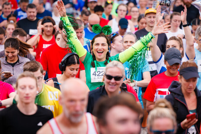 Lady in green in the middle of a crowd of runners. She has her arms outstretched and looks overjoyed to be there.
