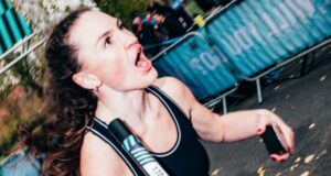 Woman wearing black vest and holding a relay baton runs past the camera. Her mouth is open in what appears to be a shout to a fellow team ember out of shot