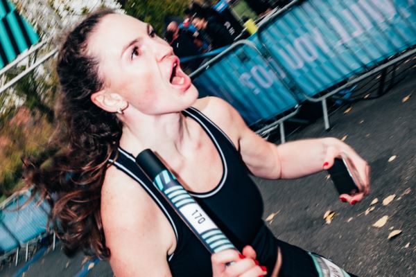 2025_18.10_ENDURE_24-1057 Woman wearing black vest and holding a relay baton runs past the camera. Her mouth is open in what appears to be a shout to a fellow team ember out of shot