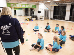 A group of primary age children wearing blue tshirts sit in a circle on a wooden floor while a lady standing next to a flipchart explains something to them